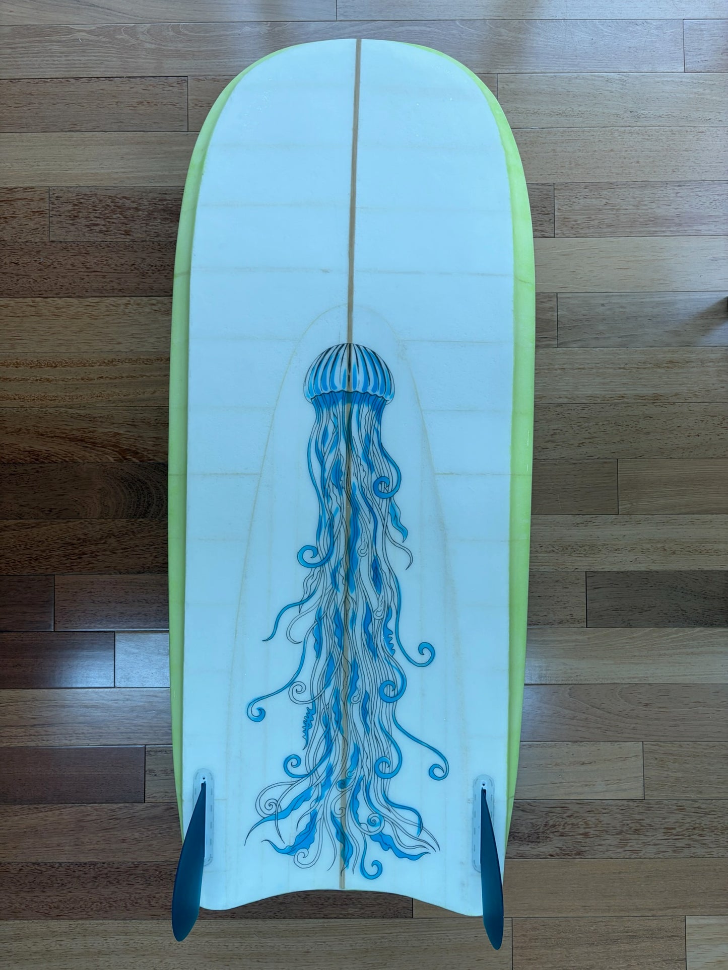 A bodyboard with a white bottom featuring a blue jellyfish graphic, bordered by a lime green edge, and two fins at each edge of the tail, resting on a wooden floor.