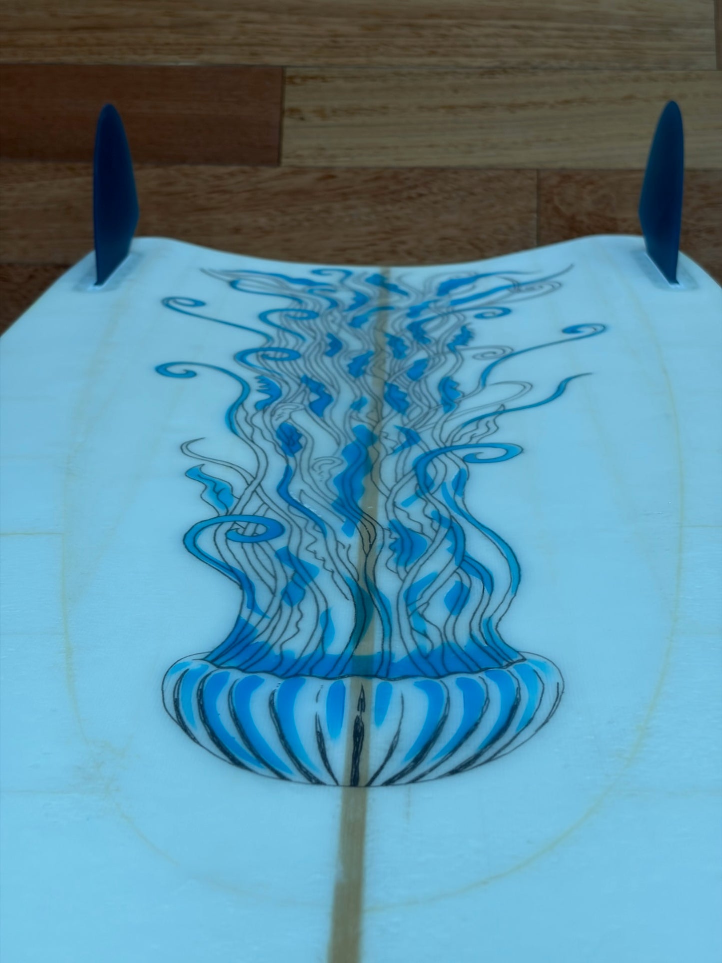 A close-up of a surfboard underside with blue fin blades and a blue jellyfish graphic running down the center on a light-colored board, photographed against a wood plank background.