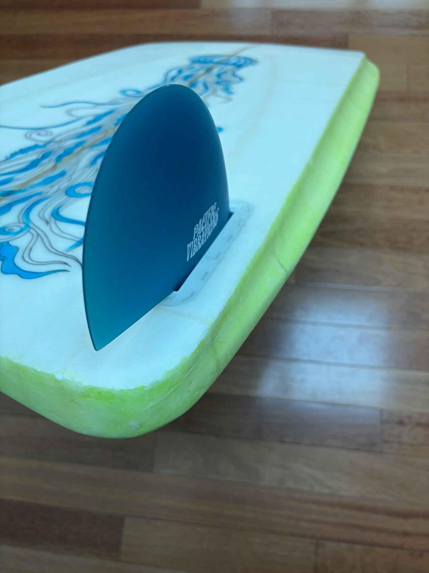A close-up of an upside-down surfboard with a bright lime green edge, a white bottom, blue jellyfish graphics, and a blue fin, resting on a wooden floor.