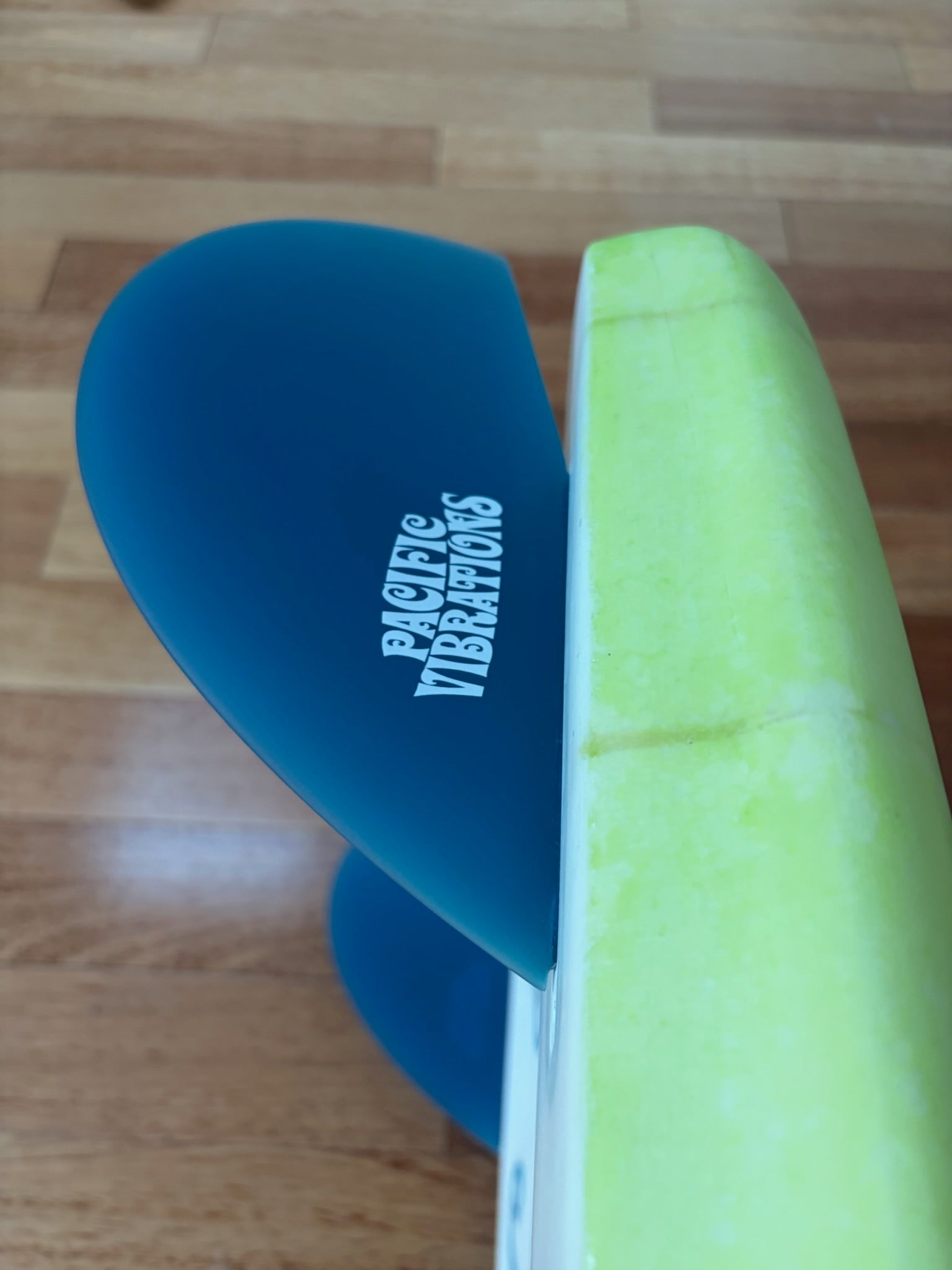 Close-up of a surfboard showing a blue fin with the white printed brand name 'Pacific Vibrations' and a bright lime-green foam rail, photographed on a wooden floor.
