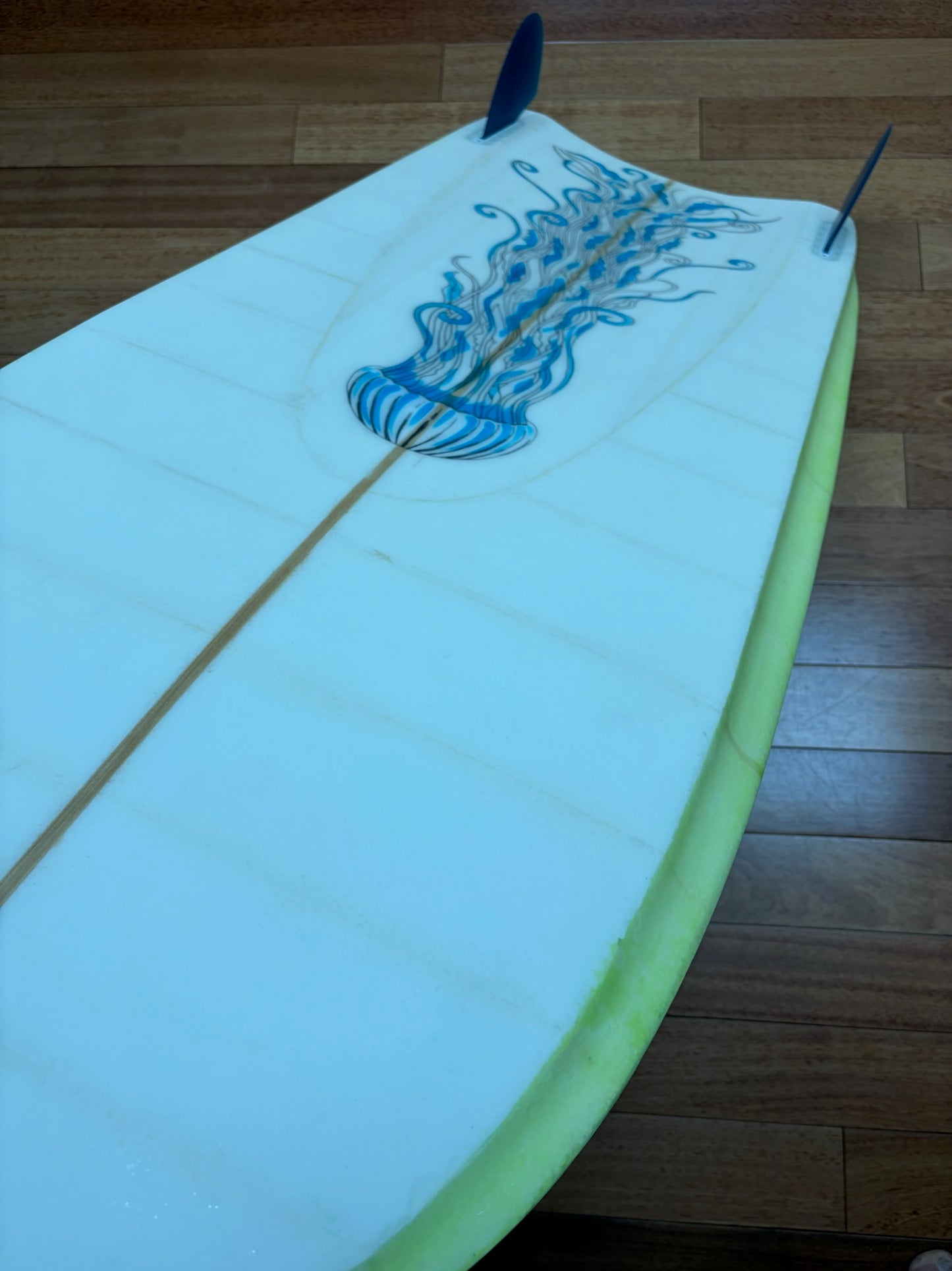 A white surfboard with a blue jellyfish graphic on the deck, resting on a wooden floor. The board has a light green edge and two blue fins.