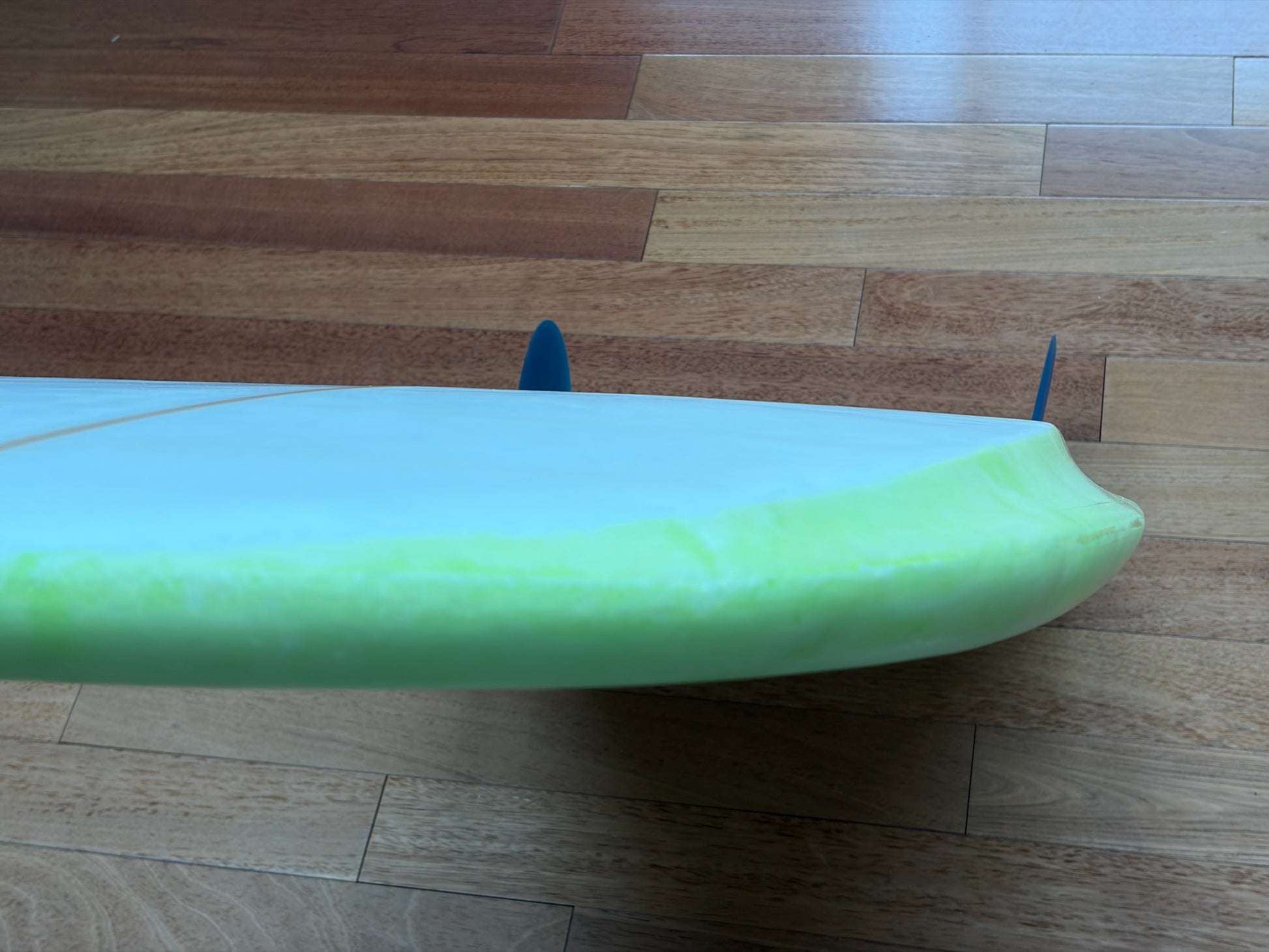 An upside-down close-up of a surfboard resting on a wooden floor, showing a white bottom surface, a bright neon green top and blue fins.