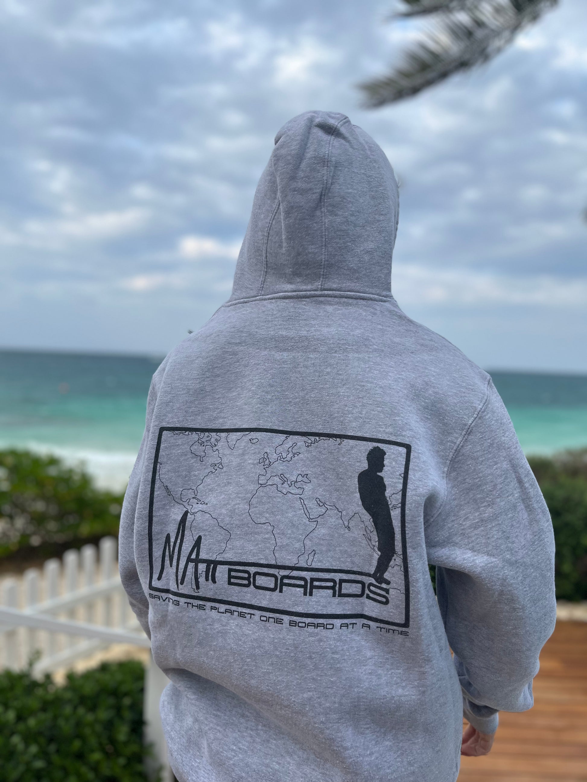 A photo of a model wearing a gray hoodie sweatshirt, taken from the back, with the hood up, at a beach setting. The sweatshirt has a design on the back of a world map with the Matt Boards logo and a saying "Saving the planet one board at a time"