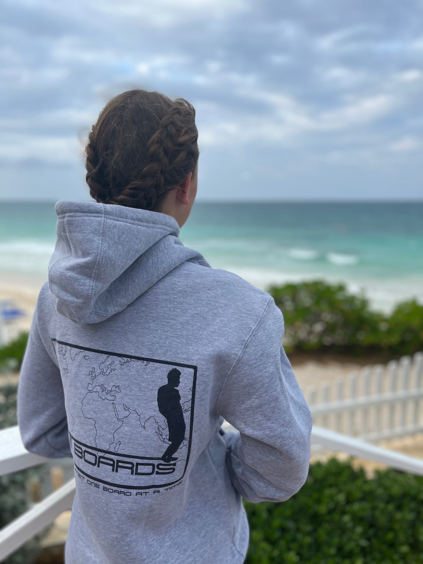 A photo of a model wearing a gray hoodie sweatshirt, taken from the back, with the hood down, at a beach setting. The sweatshirt has a design on the back of a world map with the Matt Boards logo and a saying "Saving the planet one board at a time"