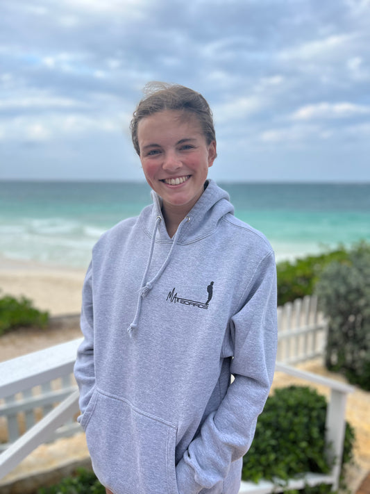 A photo of a smiling model wearing a gray hoodie sweatshirt, taken from the front, with the hood down, at a beach setting. The sweatshirt has has the Matt Boards logo on the left chest.