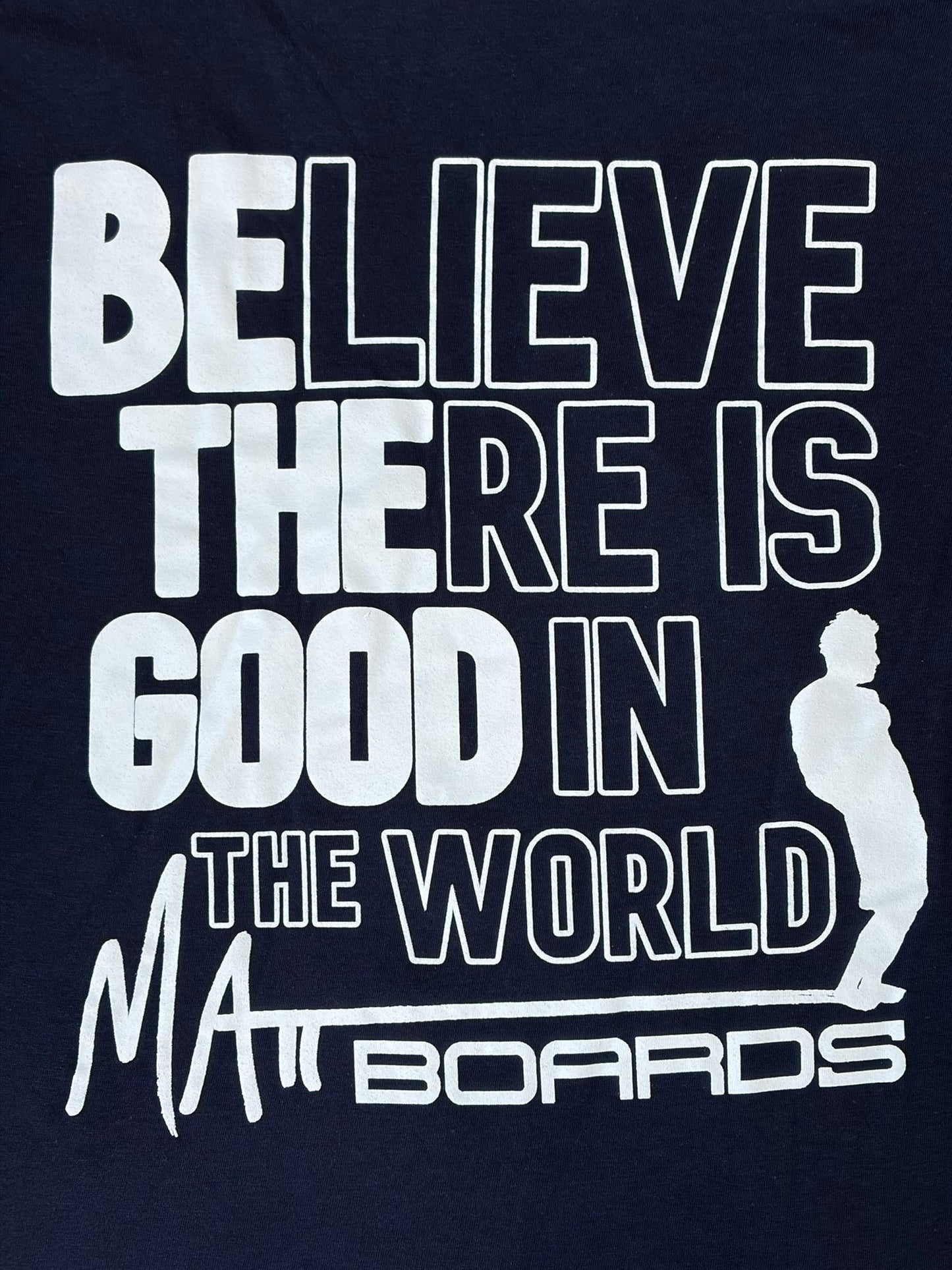 Navy blue shirt color with white motivational quote "Believe there is good in the world" with "Be the good" bolded, and a Matt Boards logo with the silhouette of a person on a surf board.