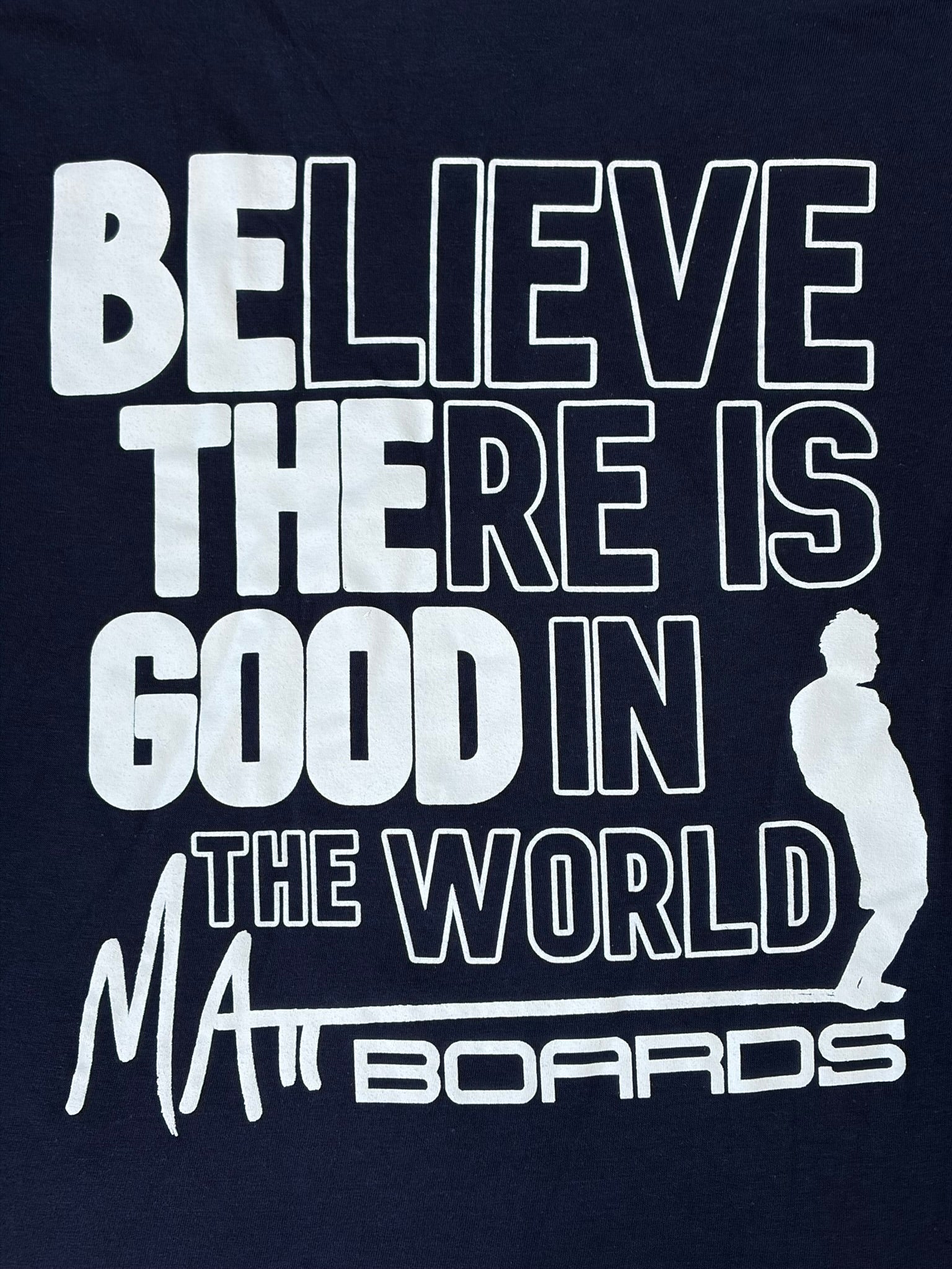 Navy blue shirt color with white motivational quote "Believe there is good in the world" with "Be the good" bolded, and a Matt Boards logo with the silhouette of a person on a surf board.