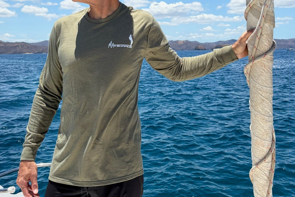 Man with a olive green long sleeve shirt with a Matt Boards logo, on a sailboat in open water with clear blue sky