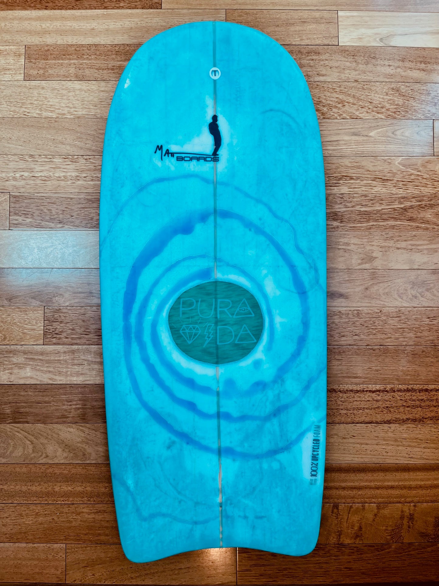 Turquoise bodyboard with a black Matt Boards logo, a swirling blue design surrounding a green circle that says Pura Vida in the middle, on a wooden background