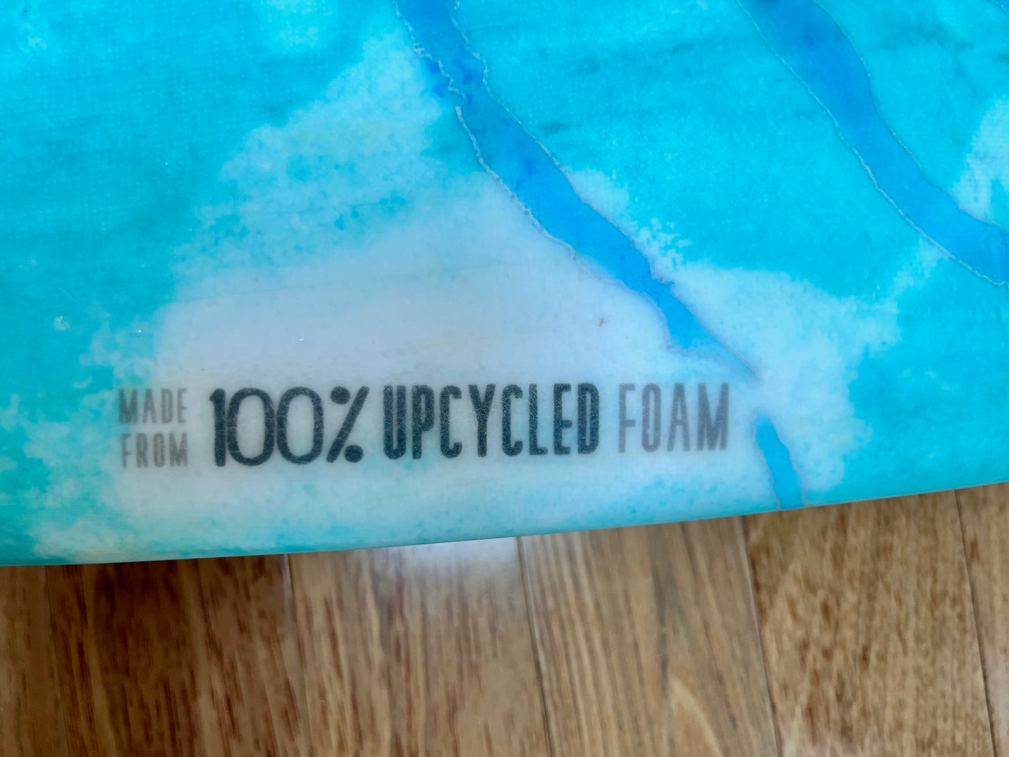 Detail of bodyboard with a label that says 'Made from 100% Upcycled Foam' with turquoise and blue color on a wood background.
