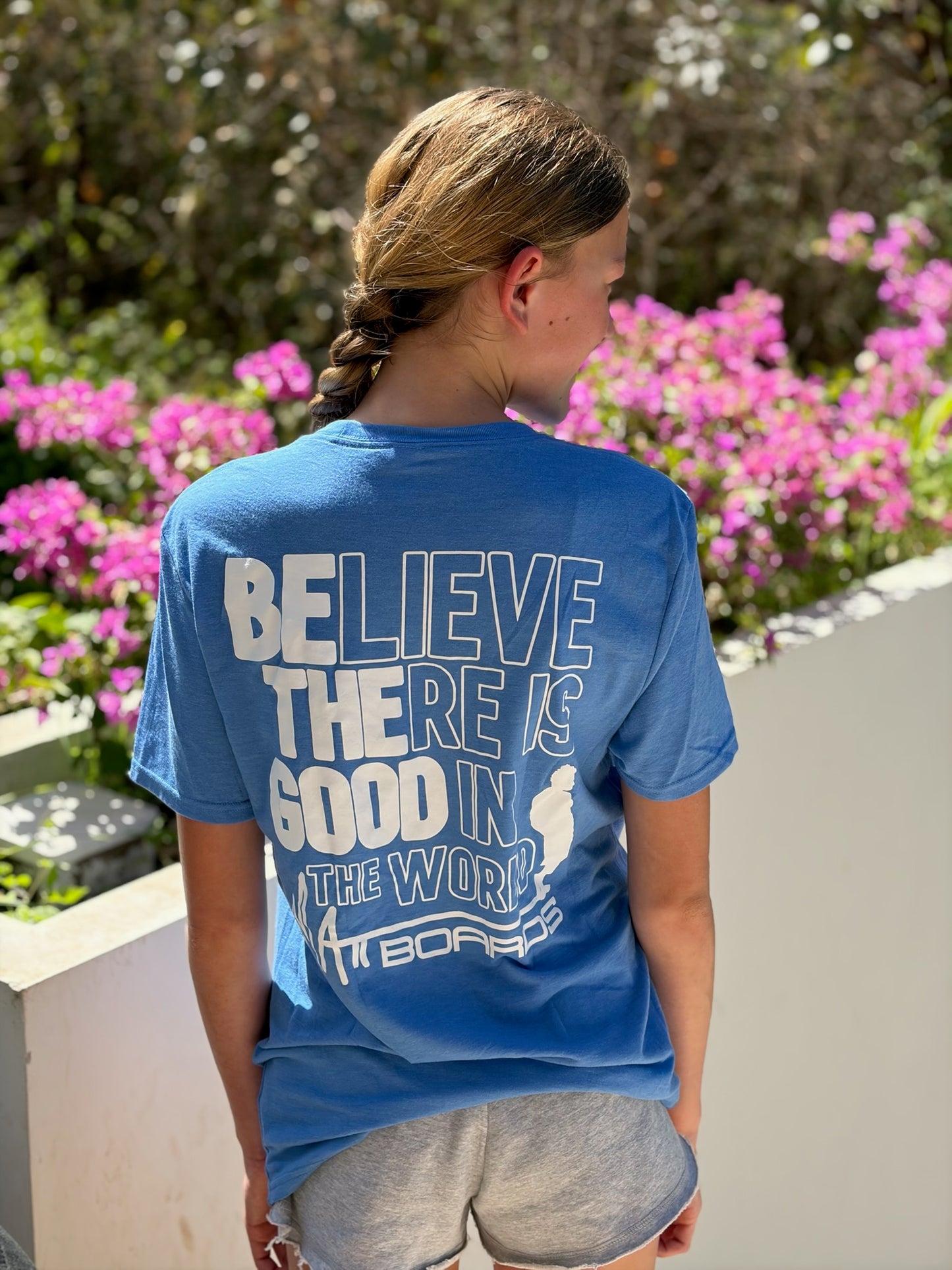 Girl wearing a blue t-shirt with a motivational quote, including believe there is good in the world with be the good bolded, on a beach