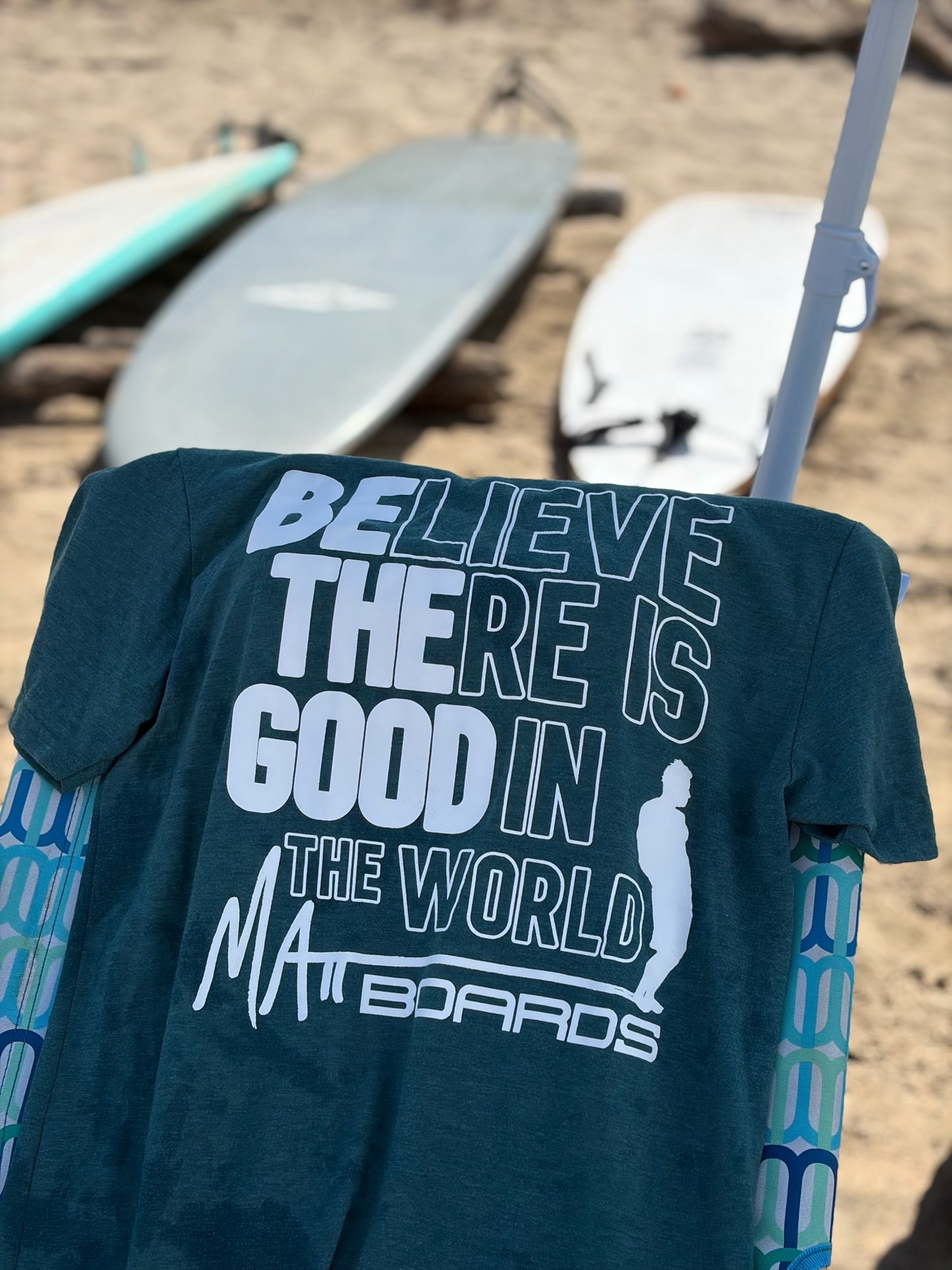 Dark green t-shirt with motivational quote on a stand with surfboards in the background