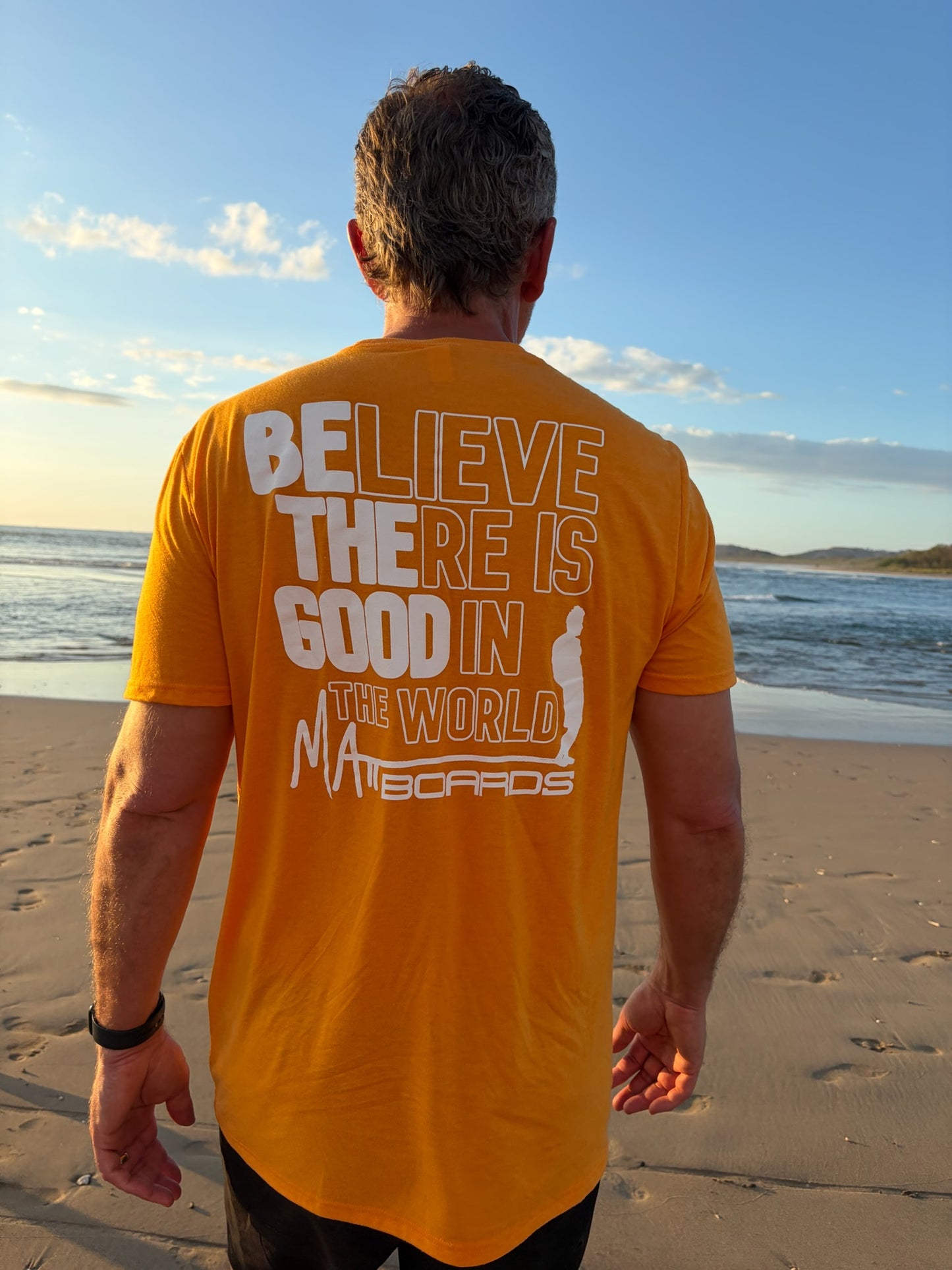 Man wearing a orange t-shirt with a motivational quote, including believe there is good in the world with be the good bolded, on a beach