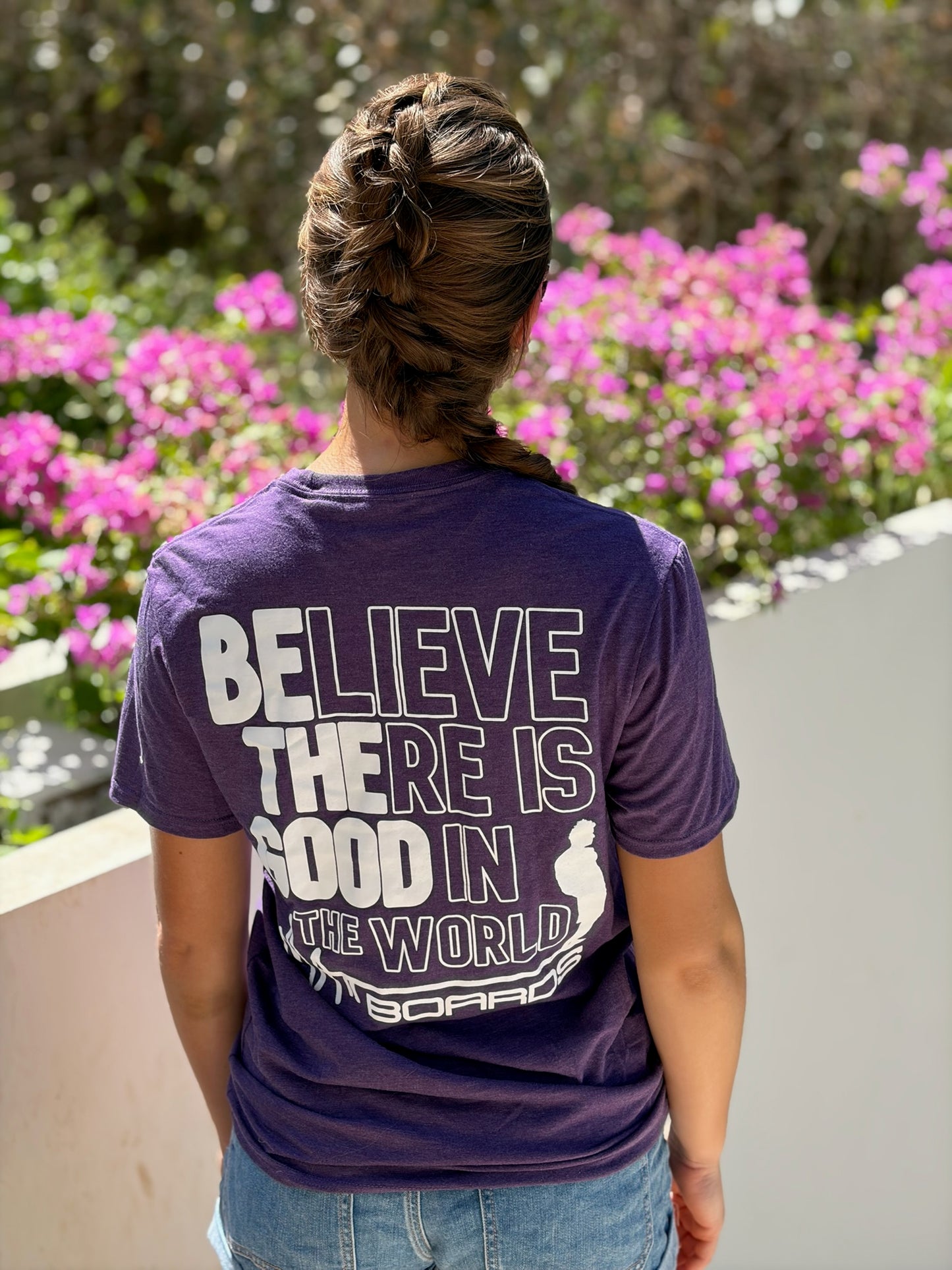 Person wearing a purple t-shirt with a motivational quote, standing in front of pink flowers.